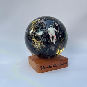 Puede incluir: Una esfera decorativa con una figura de astronauta en miniatura dentro de una resina oscura similar a una galaxia. La esfera descansa sobre una base de madera grabada con las palabras "You Are My Universe". La resina contiene detalles de pan de oro.