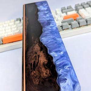 Könnte beinhalten: Handballenauflage für eine Tastatur mit dunklem Holzmaserungsdesign und einem blauen und weißen Harzwellenmuster. Mit Kupferakzent und grauer Basis. Ca. 30 cm lang.