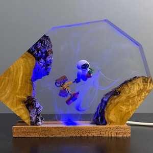May include: A unique resin lamp featuring a clear, geometric design with a blue glow. The lamp incorporates wood and a miniature robot figure. The base is made of wood, and the lamp is plugged in with a white cord.