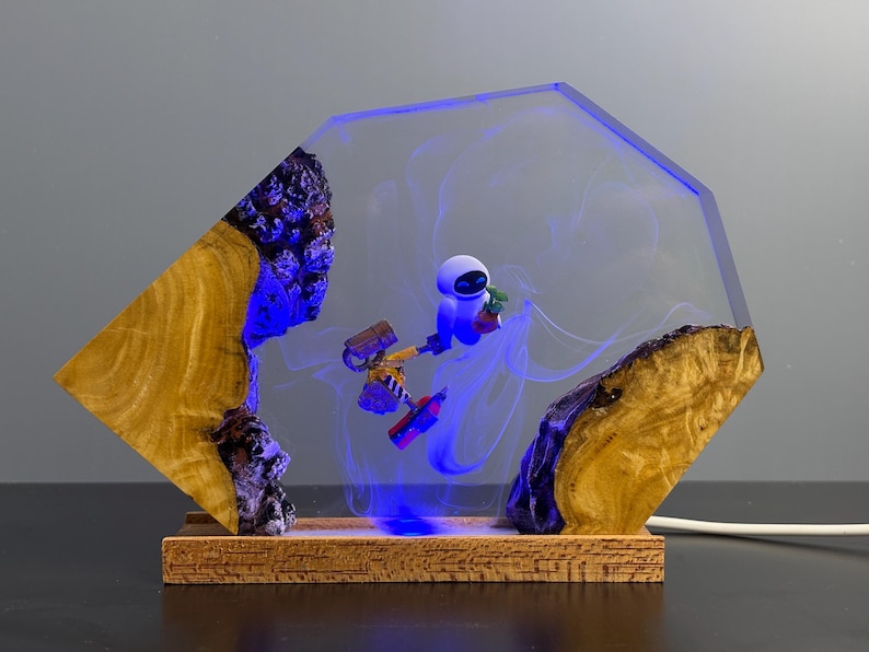 May include: A unique resin lamp featuring a clear, geometric design with a blue glow. The lamp incorporates wood and a miniature robot figure. The base is made of wood, and the lamp is plugged in with a white cord.
