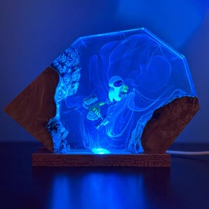 May include: A unique, handmade lamp featuring a blue-glowing astronaut inside a clear resin structure. The resin is encased in a wooden frame, creating a striking contrast. The lamp emits a soft, blue light, perfect for a nightstand or desk.