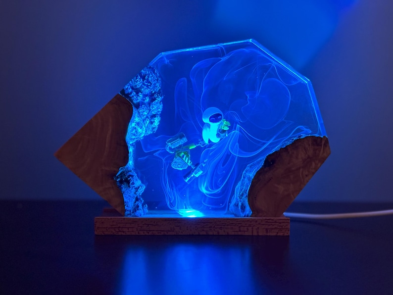 May include: A unique, handmade lamp featuring a blue-glowing astronaut inside a clear resin structure. The resin is encased in a wooden frame, creating a striking contrast. The lamp emits a soft, blue light, perfect for a nightstand or desk.