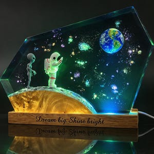 Może przedstawiać: Prostokątna lampa z żywicy z drewnianą podstawą z napisem "Dream big. Shine bright.". Część z żywicy zawiera miniaturowego astronautę i kosmitę stojących na zakrzywionej drewnianej powierzchni, z wbudowaną świecącą Ziemią i gwiazdami.