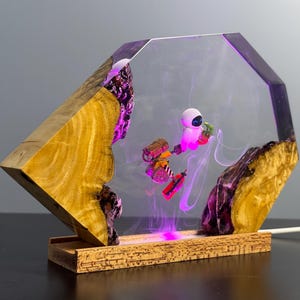May include: A unique lamp featuring a geometric wooden base and a clear resin body. Inside, a scene with animated characters is illuminated by purple and pink LED lights. The lamp's base is made of cork.