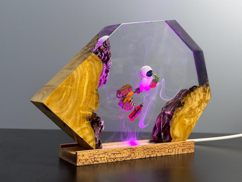 May include: A unique lamp featuring a geometric wooden base and a clear resin body. Inside, a scene with animated characters is illuminated by purple and pink LED lights. The lamp's base is made of cork.