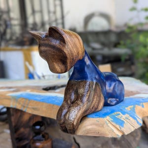 May include: A wooden cat sculpture with a smooth, polished finish. The cat's head and front legs are a warm brown, while the body is a vibrant blue. The sculpture is resting on a wooden surface with blue accents, showcasing a blend of natural and artistic elements.