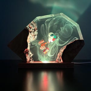 May include: A unique lamp featuring a clear, geometric resin encasing a stylized robot figure. The lamp has a wooden base and a rough, natural edge. The interior glows with a soft, ethereal light, creating a whimsical and artistic home decor piece.