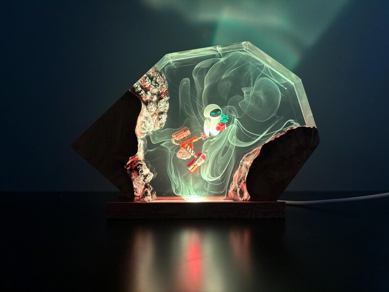 May include: A unique lamp featuring a clear, geometric resin encasing a stylized robot figure. The lamp has a wooden base and a rough, natural edge. The interior glows with a soft, ethereal light, creating a whimsical and artistic home decor piece.