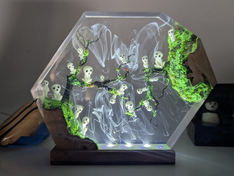 Pu&ograve; includere: Una lampada in resina esagonale con un albero bianco luminoso e piccole figure bianche. L'albero &egrave; circondato da muschio verde e la base &egrave; in legno scuro.