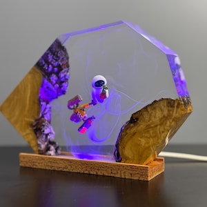 May include: A unique lamp featuring a clear, geometric resin design with a miniature astronaut figure. The lamp incorporates natural wood elements and a purple LED light, creating a striking visual effect. The base is made of wood.