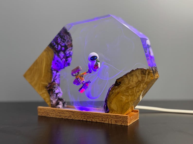 May include: A unique lamp featuring a clear, geometric resin design with a miniature astronaut figure. The lamp incorporates natural wood elements and a purple LED light, creating a striking visual effect. The base is made of wood.