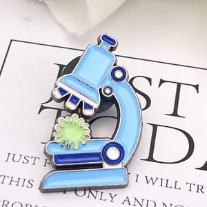 Puede incluir: Un pin de esmalte de microscopio azul claro con una célula estilizada verde y blanca. El pin tiene un contorno azul oscuro y está sobre un fondo blanco con texto negro que dice "JUST F THIS I WILL TRY TO ONLY AND NOT TACKLE PROBLEM AT ONCE".