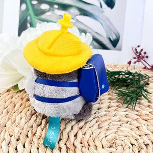 May include: A gray plush toy wearing a yellow hat and a blue backpack. The toy has a blue strap across its chest and a tag that says "2nd Grade".
