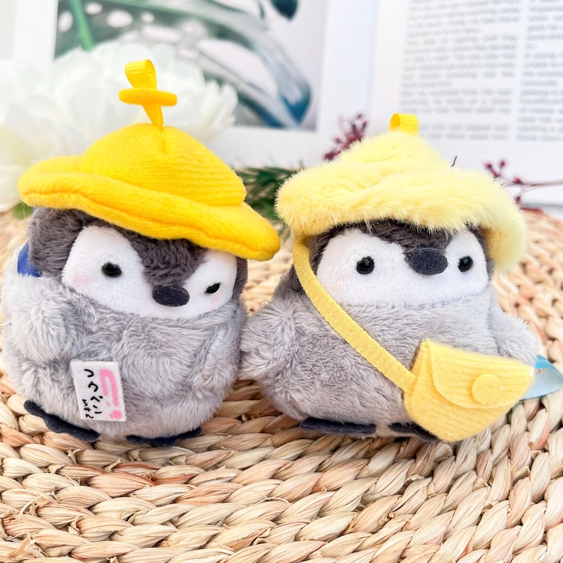 May include: Two gray penguin plush toys wearing yellow hats. One penguin has a yellow bucket hat and the other has a yellow rain hat with a strap and a small yellow bag. The penguin with the bucket hat has a pink tag with Japanese text.