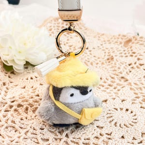 May include: A gray and white penguin plush keychain wearing a yellow hat and carrying a yellow bag. The keychain is attached to a gold ring and a white strap.
