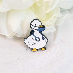 Könnte beinhalten: Ein weißer Enten-Anstecker mit einer blauen medizinischen Maske und einem Stethoskop. Die Ente hat gelbe Füße und einen schwarzen Umriss. Der Anstecker befindet sich vor einem weichen, weißen Hintergrund mit floralem Akzent.