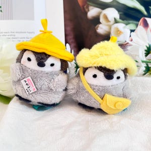 Könnte beinhalten: Zwei graue Plüschpinguine mit gelben Hüten. Ein Pinguin trägt einen gelben Hut mit einem rosa Etikett, auf dem "ペンギン" steht. Der andere Pinguin trägt einen gelben Hut mit einem gelben Band und einer gelben Tasche.