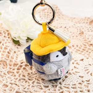 May include: A plush penguin keychain wearing a yellow hat and a blue backpack. The penguin is holding a small pink note that says "いってらっしゃい!" in Japanese.