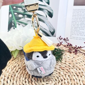 May include: A gray and white penguin plush keychain wearing a yellow hat with a white tassel. The penguin is holding a small white note that says "Go! Go! Go!" in Japanese.