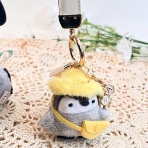 May include: A gray and white penguin plush keychain with a yellow hat and a yellow bag. The keychain has a gold chain and a gold charm with the letter "A" on it.