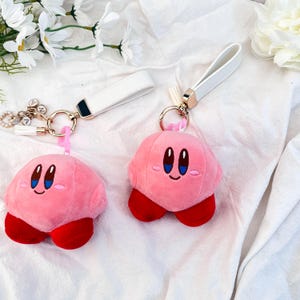 Könnte beinhalten: Zwei rosa Plüsch-Schlüsselanhänger mit einer Comicfigur mit rosa Körper, roten Füßen und blauem Gesicht. Die Schlüsselanhänger sind an weißen Lederbändern mit goldenen Ringen befestigt.
