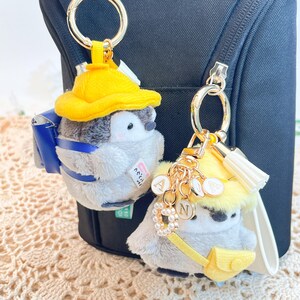 May include: Two plush penguin keychains with yellow hats and backpacks. The penguins are wearing blue and yellow backpacks. The keychain on the left has a blue backpack with the text "Kicchil" on it. The keychain on the right has a yellow backpack with a gold charm with the letter "N" on it.