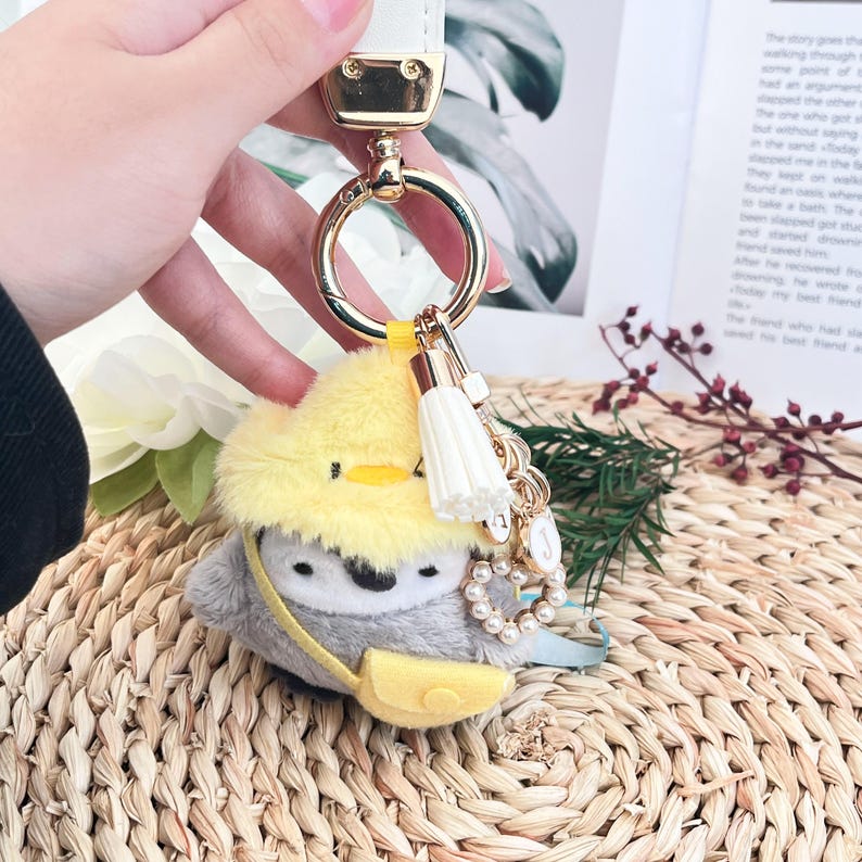 May include: A plush penguin keychain with a yellow hat and a yellow bag. The keychain has a gold chain with pearl beads and charms.