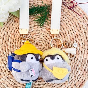 May include: Two gray penguin plush keychains with yellow hats and backpacks. One penguin has a blue backpack and the other has a yellow backpack. Both penguins have a white strap attached to their backpacks. The keychains are attached to white leather straps with gold hardware.