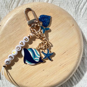 Könnte beinhalten: Ein goldener Schlüsselanhänger mit einem blauen Wal-Anhänger, einem blauen Seestern-Anhänger, einem blauen Glitzerherz-Anhänger und einem weißen Perlenanhänger mit dem Wort "GRACE" in goldenen Buchstaben.