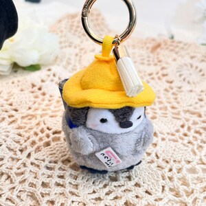 May include: A gray and white penguin plush keychain wearing a yellow hat with a white tassel. The penguin is holding a small blue and white sign that says "こんにちは".
