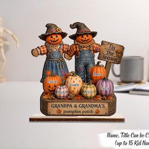 May include: A decorative tabletop display featuring two scarecrows with jack-o'-lantern heads wearing hats and overalls. A wooden sign reads 'Tis the season' and 'GRANDPA & GRANDMA'S pumpkin patch'. Several small pumpkins are labelled with names.