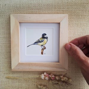 以下が含まれることがあります： 枝にとまったシジュウカラを描いた小さな絵画が入った正方形の木製フレーム。鳥は黒い頭、白い頬の斑点、黄色い胸、灰色がかった青い翼を持ち、白い背景に描かれています。