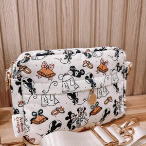 May include: A white rectangular crossbody bag featuring a repeating pattern of wedding-themed icons including bells, rings, and cartoon mice characters. The bag has gold-tone hardware, a front zipper pocket with a circular charm, and a detachable strap.