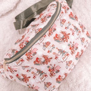 May include: A light pink fanny pack with a repeating pattern of Christmas-themed illustrations. The illustrations include children, reindeer, and toys. The bag has a dark green adjustable strap and a gold zipper.