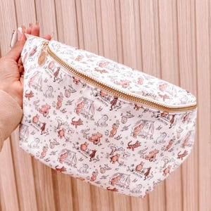 May include: White fanny pack with a repeating pattern of cartoon animals in pink and brown. The bag has a gold zipper and a small gold tag. The fanny pack is a practical accessory for carrying essentials.