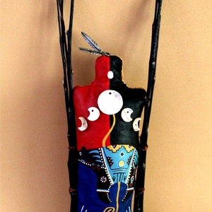 Puede incluir: Una muñeca artesanal nativa americana con un diseño pintado en rojo, negro y azul. La muñeca está adornada con círculos blancos y lunas crecientes. La muñeca está de pie sobre un manojo de palos.