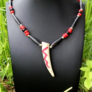 Puede incluir: Un collar artesanal con un colgante en forma de diente decorado con un diseño de zigzag rojo. El collar tiene cuentas negras, rojas y plateadas. El colgante es de color crema y las cuentas están ensartadas en un cordón negro. El collar se exhibe en un soporte negro.