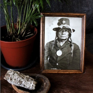 Puede incluir: Una fotografía en blanco y negro enmarcada de un hombre nativo americano que lleva un sombrero con una estrella. Lleva una chaqueta oscura y tiene una trenza larga. La foto está en un marco de madera con un paspartú marrón.