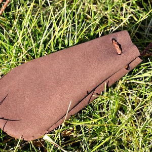 Puede incluir: Una bolsa de cuero marrón con cierre de cordón. La bolsa está colgada de un cordón trenzado marrón.