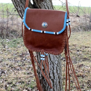 Puede incluir: Un bolso de cuero marrón cruzado con flecos. El bolso tiene un ribete de cuentas azules y blancas y un concho plateado.