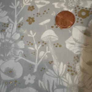 May include: A close-up of a fabric with a floral pattern in white and grey, accented with gold flecks. A copper penny is on the fabric, providing a size reference. The design includes stylized flowers, mushrooms, and foliage.