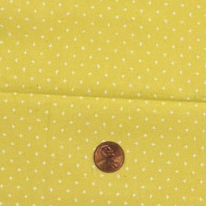 Puede incluir: Una tela de color amarillo brillante con un patrón repetitivo de pequeñas estrellas blancas de cuatro puntas. Una moneda de color cobre está colocada sobre la tela para proporcionar una escala del tamaño del estampado.