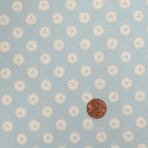 May include: A light blue fabric featuring a repeating pattern of small white circular motifs, each containing a tiny three-leaf clover design. A copper-colored United States penny is placed on the fabric to provide a sense of scale.
