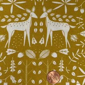 May include: Mustard-colored fabric with a repeating pattern of white deer, leaves, and floral elements. A penny is included for scale. The design features two deer facing each other, surrounded by stylized foliage. The fabric has a textured appearance.