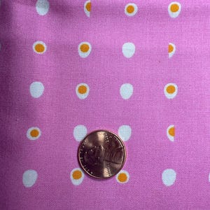 Puede incluir: Tela rosa con un patrón repetido de círculos blancos y diseños en forma de huevo naranja y blanco. Se coloca una moneda de cobre en la tela para la escala.