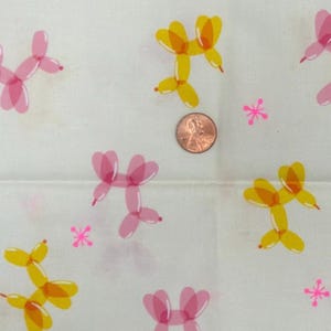 May include: White fabric featuring a repeating pattern of pink and yellow balloon animals and pink starburst shapes. A penny is included for scale. The balloons are in the shape of dogs. The fabric is likely cotton or a similar material.
