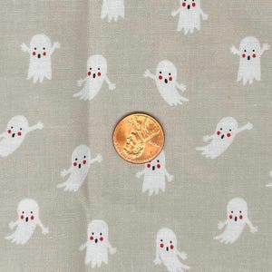 May include: A close-up of a light grey fabric featuring a pattern of white cartoon ghosts. Each ghost has red cheeks and a mouth. A penny is in the centre for scale. The fabric is suitable for a Halloween-themed craft project.