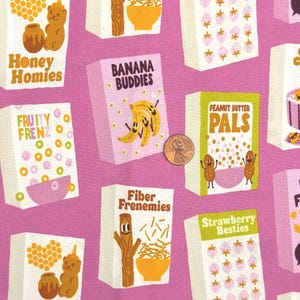 Könnte beinhalten: Ein rosa Stoff mit einem wiederkehrenden Muster aus Müslischachteln, die verschiedene Zeichentrickfiguren und Texte wie Honey Homies, Banana Buddies, Peanut Butter Pals und Fiber Frenemies zeigen. Eine Kupfermünze dient als Größenvergleich.