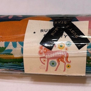 Puede incluir: Un paquete enrollado de tiras de tela envuelto en plástico transparente con una etiqueta de papel. La etiqueta presenta una ilustración de un caballo de carrusel rosa, el texto "RUBY STAR SOCIETY" y "CAROUSEL BY MELODY MILLER", atado con una cinta negra impresa con "RUBY STAR".