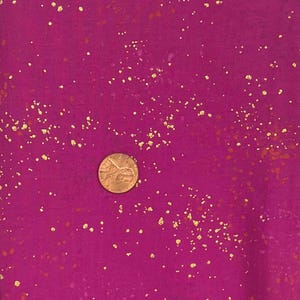 以下が含まれることがあります： 表面に金色の斑点が散りばめられたマゼンタ色の生地のクローズアップ。生地の上に硬貨が置かれ、金色のアクセントの質感とサイズを示しています。生地は柔らかくマットな仕上がりです。